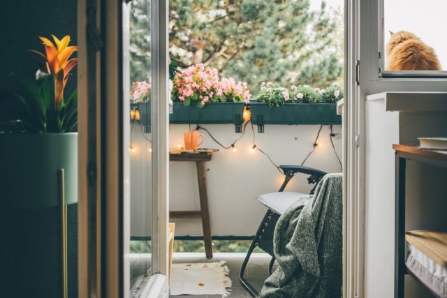 An open patio door reveals an apartment balcony with a chair, side table, and string lights.