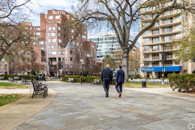 Two businessmen walking on a path through a park with apartment buildings in the background.