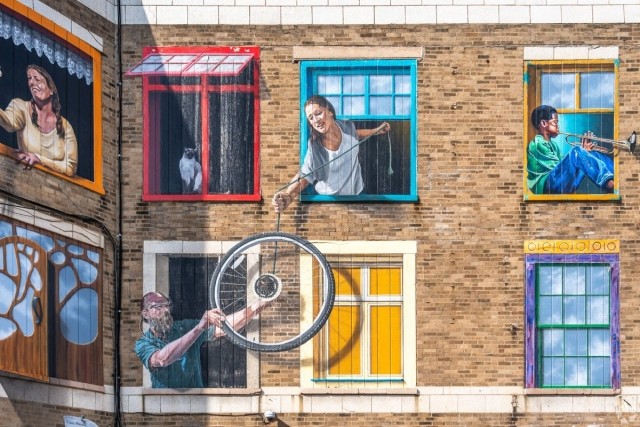 A vibrant mural on a brick building featuring multiple painted window scenes, including a white cat in a red frame, a woman reaching out from a blue frame with a rope, a boy in a yellow frame playing a trumpet, and a man holding a bicycle wheel from a lower window.