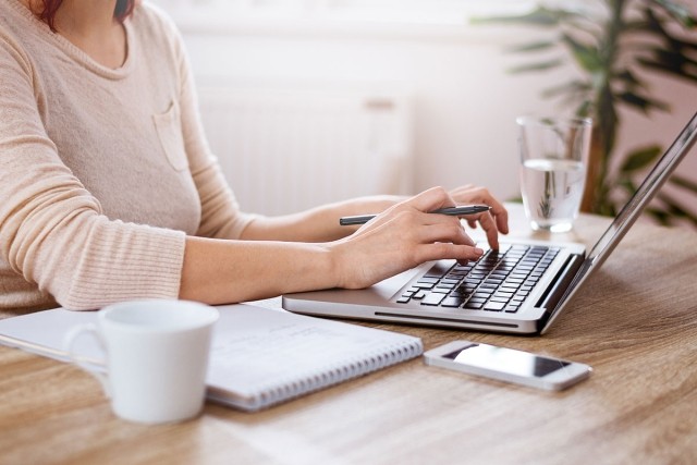Woman holding pen and typing on laptop with notebook and phone next to her.
