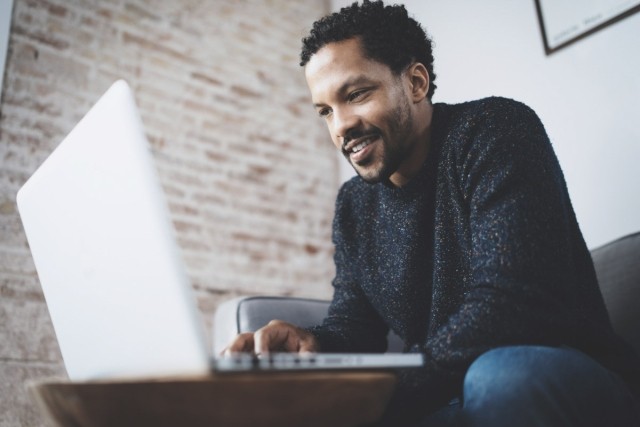 A smiling young man searches for apartments on a laptop.