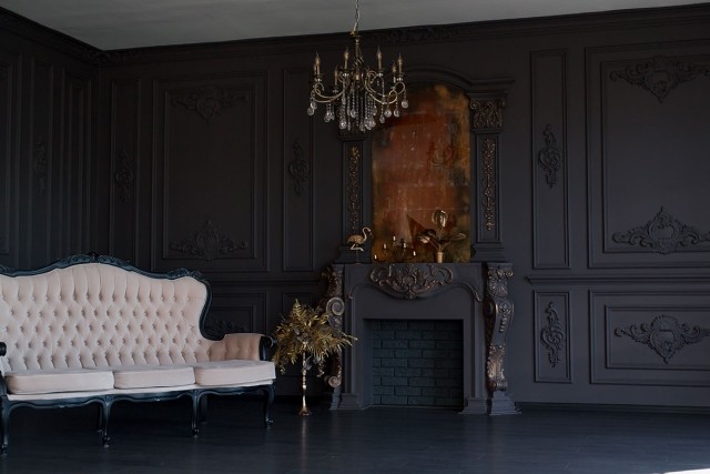 An opulent living room showcases paneled walls, a chandelier, a carved fireplace that are all black as well as a pale upholstered sofa in low light.