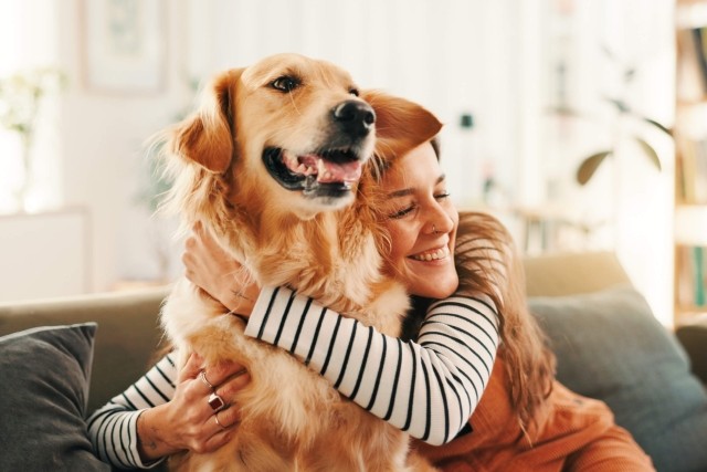 A woman with long brown hair in a striped shirt and orange overalls hugs her Golden Retriever.