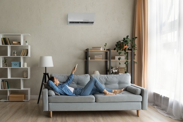 A woman in a denim jacket and jeans lies on her couch in her living room. She is adjusting her AC unit with a remote control.