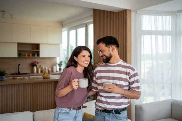 A woman and a man drink coffee while standing in their spacious living room. They are smiling and have their arms around each other.