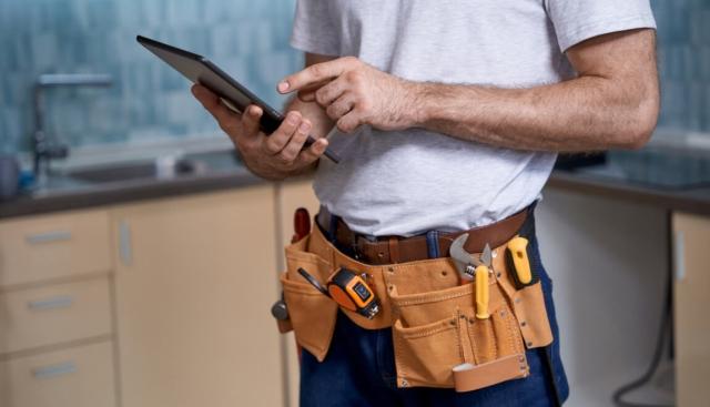 A man wearing a tool belt checks a tablet for maintenance requests.