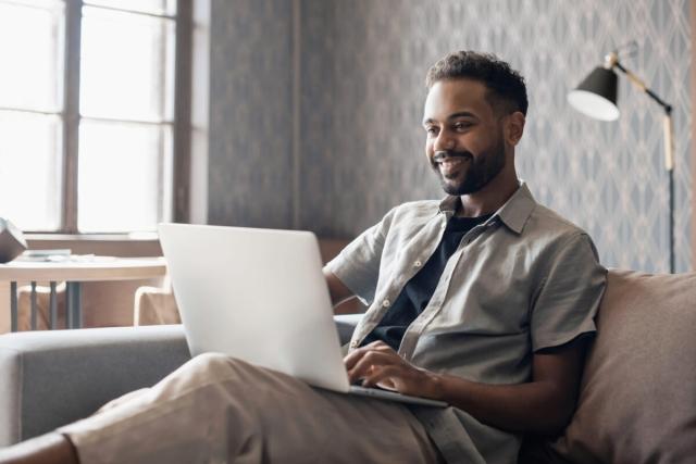A young man browses on his laptop while sitting on a couch.