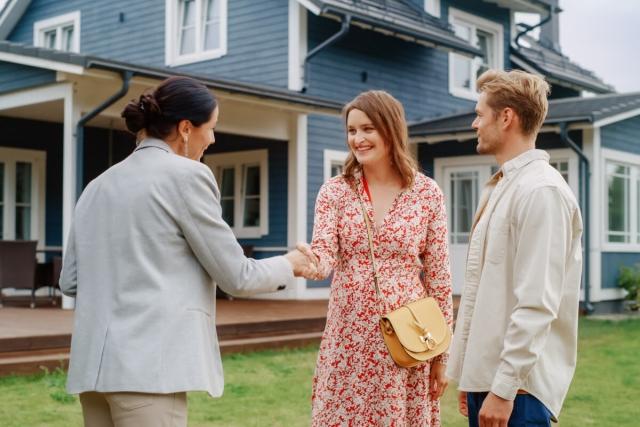 A young couple shakes hands with a landlord in front of a blue rental house.