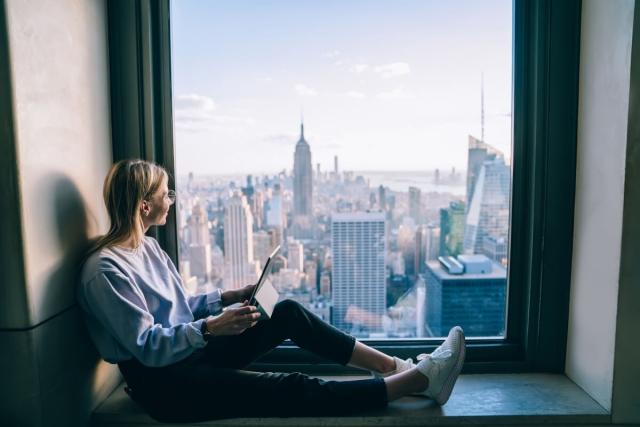 A young woman sits by a window looking at the New York City skyline.