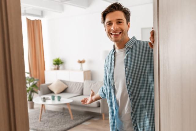 A man smiles at the camera and welcomes an unseen guest into his apartment.