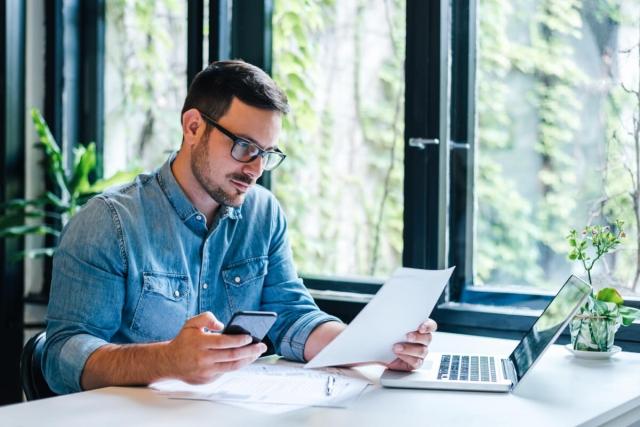 A man with glasses studies a piece of paper with his laptop open next to him.