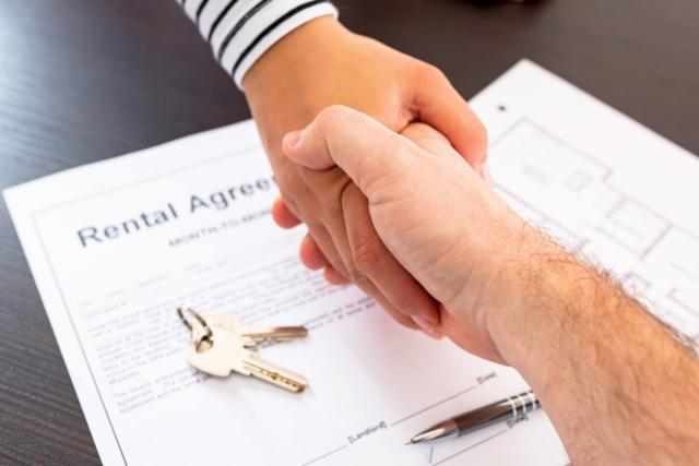 A landlord and renter shake hands over a rental agreement and a set of keys.