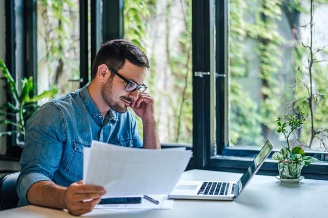 Man with glasses sitting at desk and reading on laptop screen as he holds papers.