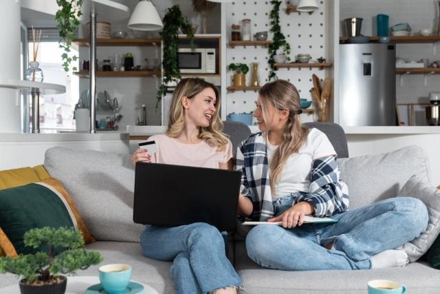 Two young women sharing a modern apartment living room, shopping online together with a laptop and credit card, ideal for roommates splitting rent in a stylish rental space.