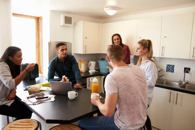 College students enjoying breakfast in a shared apartment kitchen with modern white cabinetry and communal dining space.
