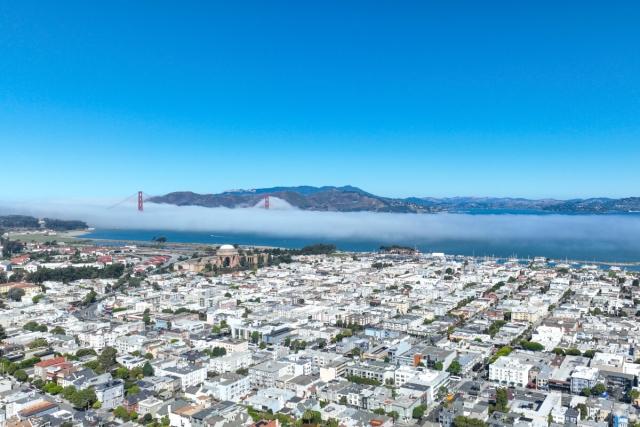 An aerial view of a San Francisco neighborhood shows the Golden Gate Bridge and a mountain range in the background.
