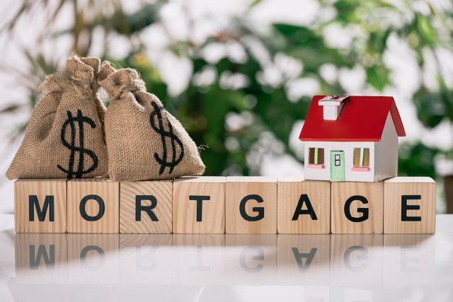 The word "mortgage" spelled out on wooden block with a model house and bags of money on top of it.