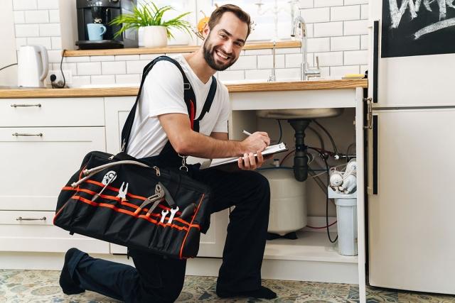Man kneeling in front of sink he is fixing and smiling while he writes.