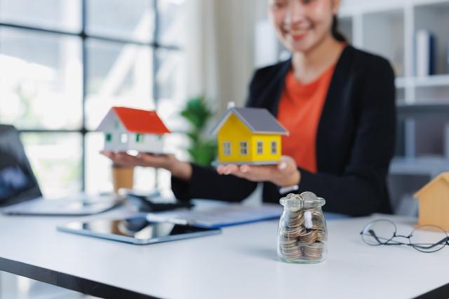 Woman holding two model houses in her hands with a jar of coins in front, representing comparing properties.
