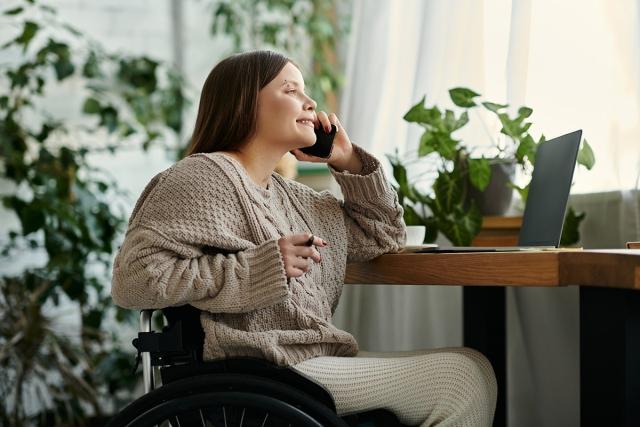 Woman in wheelchair smiles as she is on the phone at her desk.