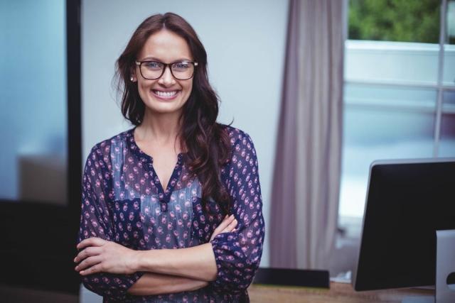 A smiling business lady leaning against a desk with arms folded