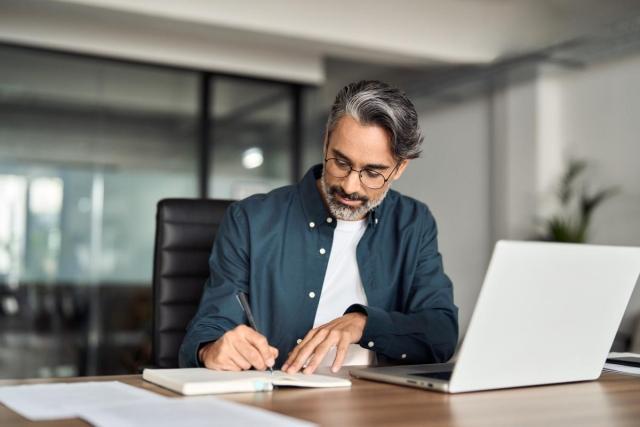 A man sitting in front of his laptop and writing in a notepad