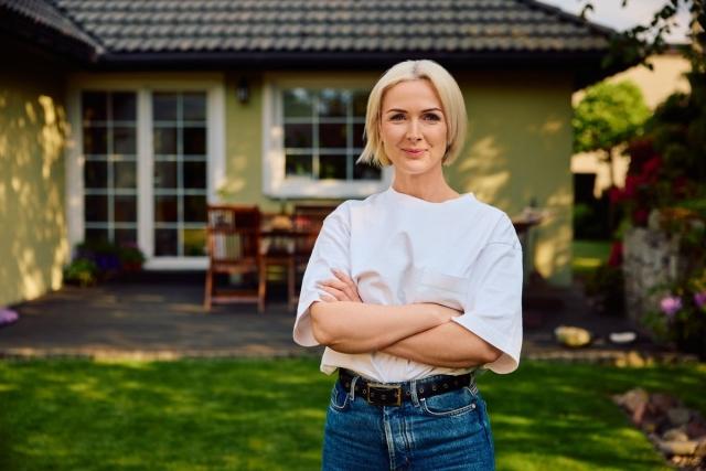 A confident woman standing outside of a small home with arms folded..