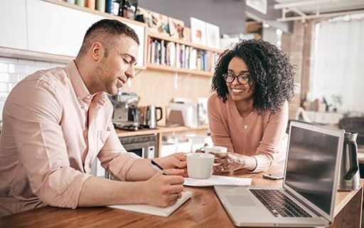 a couple conversing while seated at a table with coffee, laptop, and notepad