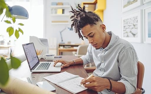 person sitting at a desk in a cozy office with their laptop open and reviewing an accounting ledger