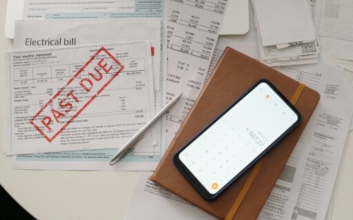 A pile of utility bills marked with a bright red "PAST DUE" stamp sits next to a phone with a calculator app open.