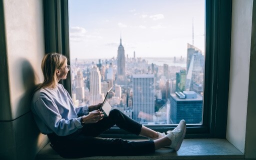 A young woman sits by a window looking at the New York City skyline.