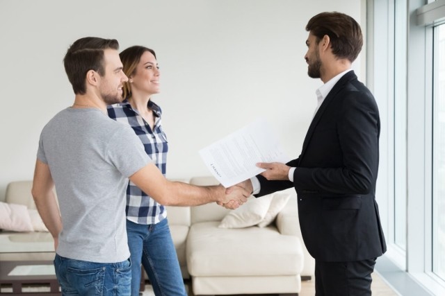 A landlord wearing a suit shakes hands with a couple touring his property.