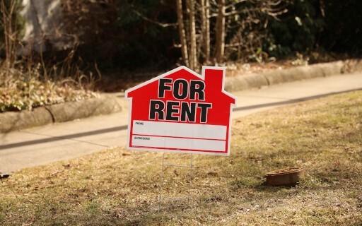 A red "For Rent" sign in the shape of a house is stuck in the grass by a sidewalk.