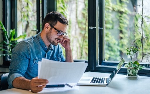 Man with glasses sitting at desk and reading on laptop screen as he holds papers.