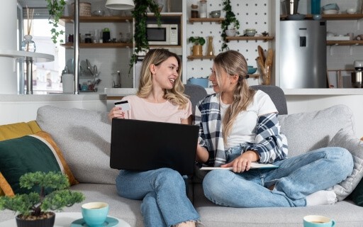 Two young women sharing a modern apartment living room, shopping online together with a laptop and credit card, ideal for roommates splitting rent in a stylish rental space.