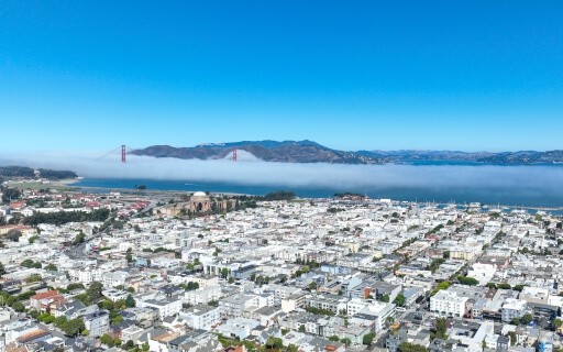 An aerial view of a San Francisco neighborhood shows the Golden Gate Bridge and a mountain range in the background.