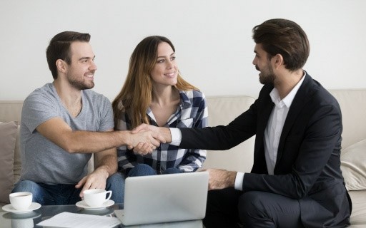 A young landlord wearing a suit shakes hands with a young couple looking to rent.