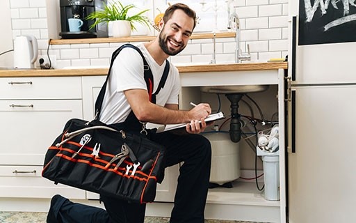 Man kneeling in front of sink he is fixing and smiling while he writes.