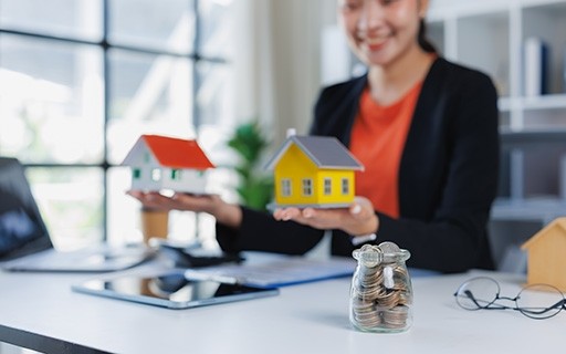 Woman holding two model houses in her hands with a jar of coins in front, representing comparing properties.