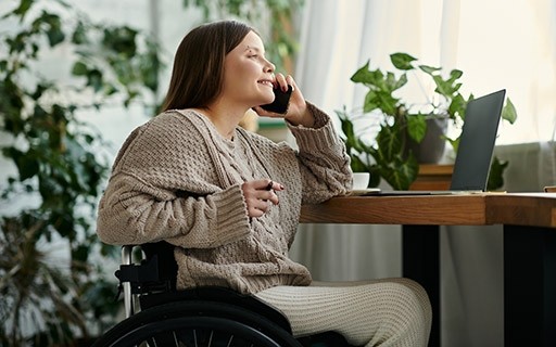 Woman in wheelchair smiles as she is on the phone at her desk.