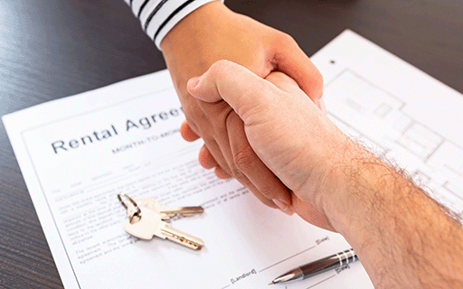 A landlord and renter shake hands over a rental agreement and a set of keys.
