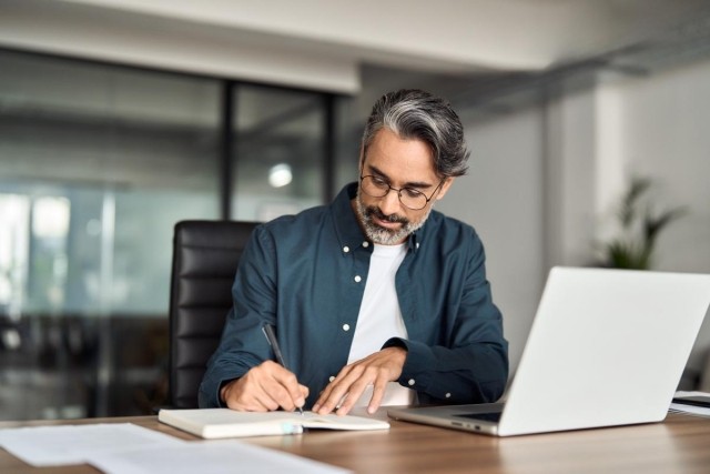 A man sitting in front of his laptop and writing in a notepad